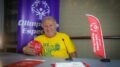 zico sentado atrás de uma mesa, segurando uma bola vermelha e sorrindo para a camera. há vários materiais e banners das Olimpiadas especiais atrás dele e sobre a mesa.