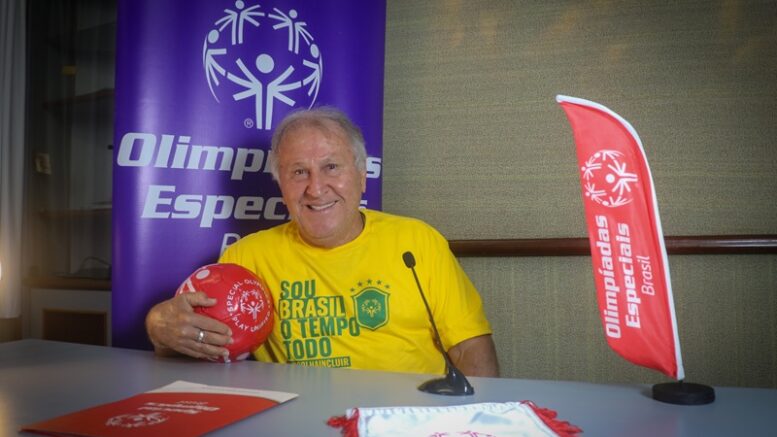 zico sentado atrás de uma mesa, segurando uma bola vermelha e sorrindo para a camera. há vários materiais e banners das Olimpiadas especiais atrás dele e sobre a mesa.