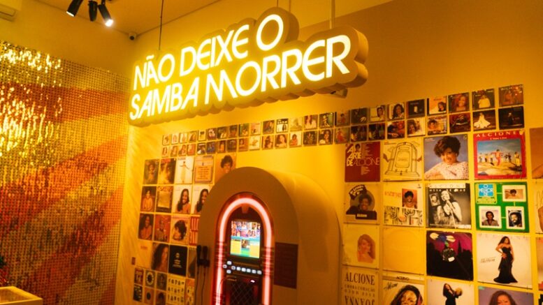 Parede em tons de amarelo com muitas fotos, uma juckbox e em neon a frase Não deixe o samba morrer