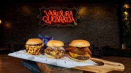 três mini hamburguers sobre uma tábua retangular, colocada sobre uma pilha de livros. No fundo, uma parede de tijolos com a logo do Vassoura Quebrada.