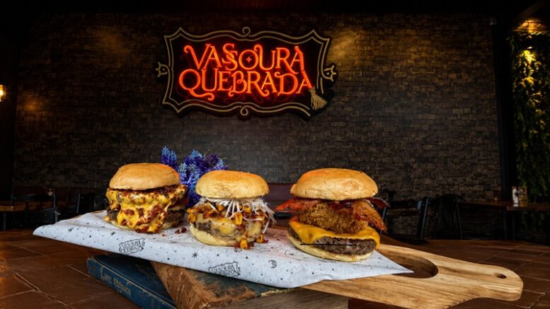 três mini hamburguers sobre uma tábua retangular, colocada sobre uma pilha de livros. No fundo, uma parede de tijolos com a logo do Vassoura Quebrada.