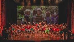 no palco com uma projeção de bonecos gigantes de frevo ao fundo, estão dezenas de bailarinos em roupas coloridas dançando frevo