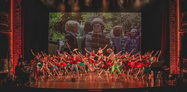 no palco com uma projeção de bonecos gigantes de frevo ao fundo, estão dezenas de bailarinos em roupas coloridas dançando frevo