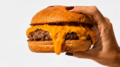 imagem de uma mão feminia segurando um amburguer de carne com queijo cheddar escorrendo na lateral