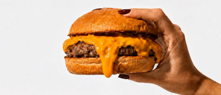 imagem de uma mão feminia segurando um amburguer de carne com queijo cheddar escorrendo na lateral