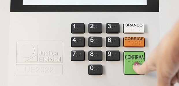 imagem do teclado da urna eletrônica com dedo feminino apertando a tecla 'confirma'.