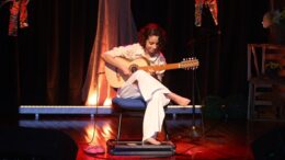 violonista Mel Moraes sentada num banco no palco, com roupas claras e descalça.