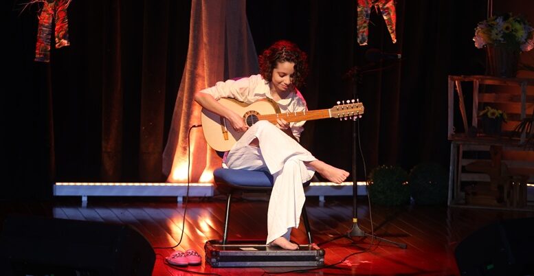 violonista Mel Moraes sentada num banco no palco, com roupas claras e descalça.